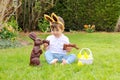 Cute Little Baby Boy With Bunny Ears On Head Holding  Chocolate Easter Bunnies Sitting On Green Grass