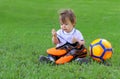 Cute little baby boy sitting with orage soccer ball on green grass holding football boots in his hands