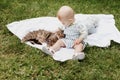 Cute little baby boy sitting on white blanket on green grass in summer, on a Sunny day, playing with a cat. Selective Royalty Free Stock Photo