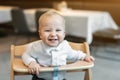 Cute little baby boy in white polo t-shirt sitting in wooden baby chair and laughing at cafe indoors. Portrait of Royalty Free Stock Photo