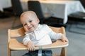 Cute little baby boy in white polo t-shirt sitting in wooden baby chair and laughing at cafe indoors. Portrait of Royalty Free Stock Photo