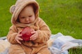 A cute little baby girl, dressed in a warm jumpsuit, is sitting on a plaid blanket on a green lawn, holding a red apple. Royalty Free Stock Photo