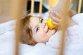 Cute little baby girl holding bottle with formula mild and drinking. Child in baby cot bed before sleeping Royalty Free Stock Photo