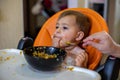 Cute little baby girl in an orange child seat eats with a hand in her mouth and looks thoughtfully. blurred spoon held