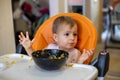 Cute little baby girl in an orange child seat in front of a plate of food throws up her hands and looks to the parent Royalty Free Stock Photo