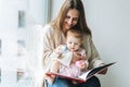 Cute little baby girl in pink dress with her mother young woman reading book in room at home Royalty Free Stock Photo