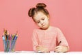 A cute little baby girl is sitting at the table and drawing with enthusiasm. Pink Background Close-Up