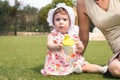 A cute little baby girl toddler wearing a white bonnet and a dress sat on green grass with her grandmother and holding a sippy cup