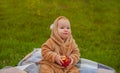 A cute little baby girl in a warm jumpsuit is sitting on a plaid on a green lawn with a red apple Royalty Free Stock Photo