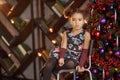 Beautiful kid girl 5-6 year old wearing stylish dress sitting in armchair over Christmas tree in room. Looking at camera. Holiday