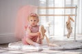 A cute little ballerina in a pink ballet costume sits near the barre in the room and tries to put on her pointe shoes. Kid and Royalty Free Stock Photo