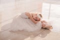 Cute little ballerina in white ballet costume and pointe shoes is posing on floor