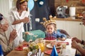 Cute little birthday girl wearing inflatable crown blowing candles on her cake, with her family around her, sitting at kitchen