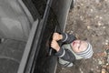 Cute little blond boy in striped hat struggles to open door of black, dusty car, hanging on handle. Family travel Royalty Free Stock Photo