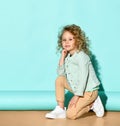 Blond girl in casual clothing sits on floor hugging knee, tilting her head and smiling. Studio portrait on turquoise background Royalty Free Stock Photo