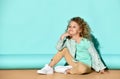 Cute blond girl in casual clothing sits on floor hugging knee, tilting her head and smiling.  portrait on turquoise background Royalty Free Stock Photo