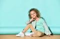 Cute blond girl in casual clothing sits on floor hugging knee, tilting her head and smiling. portrait on turquoise background Royalty Free Stock Photo