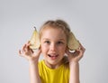 Cute little blond girl with cheerful smile peeking out from behind two halves of pear she is holding in hands. Close up