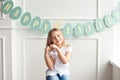 Cute little blond girl sitting in a bright room near the window on the background of streamers with the inscription