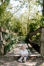 Cute Little Blond Haired Toddler Boy Kid Child Sitting and Laughing in Front of Wooden Bridge Over a Creek at the Outdoor Park in Royalty Free Stock Photo