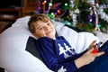 Cute little blond kid boy playing with a video game on gadget console on Christmas with decorated tree on background Royalty Free Stock Photo