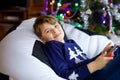 Cute little blond kid boy playing with a video game on gadget console on Christmas with decorated tree on background Royalty Free Stock Photo