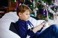 Cute little blond kid boy playing with a video game on gadget console on Christmas with decorated tree on background Royalty Free Stock Photo