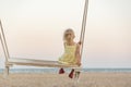 Cute little blonde girl gets on the swings on the beach on sea background. Holidays with children on the sea. Magical light of Royalty Free Stock Photo
