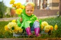 Cute little blonde girl in green raincoat sitting in a box with yellow flowers on a green lawn under rain drops or the Royalty Free Stock Photo
