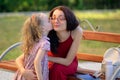 Cute Little Blonde Girl is Kissing Her Young Brunette Mother in Eyesglasses and Red Dress Sitting on the Bench in the Royalty Free Stock Photo