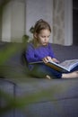 A cute little blonde girl is reading a large colorful book sitting on the sofa in the living room. Self-education of Royalty Free Stock Photo