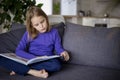 A cute little blonde girl is reading a large colorful book sitting on the sofa in the living room. Self-education of Royalty Free Stock Photo