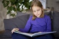 A cute little blonde girl is reading a large colorful book sitting on the sofa in the living room. Self-education of Royalty Free Stock Photo