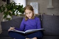 A cute little blonde girl is reading a large colorful book sitting on the sofa in the living room. Self-education of Royalty Free Stock Photo