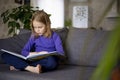 A cute little blonde girl is reading a large colorful book sitting on the sofa in the living room. Self-education of Royalty Free Stock Photo