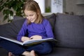 a little blonde girl is reading a big book sitting on the sofa in the living room.Smile and interest in reading.Self Royalty Free Stock Photo