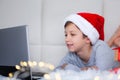 Cute little boy child in red santa hat at home looking at laptop computer screen watching online Christmas film Royalty Free Stock Photo