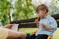 Cute little boy eating chopped carrots from lunch box, sitting on a park bench.