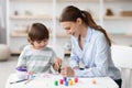 Cute little boy enjoying art classes, drawing picture with colorful paints, sitting at desk with teacher and laughing Royalty Free Stock Photo