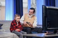 Cute little boy with grandfather sitting on sofa and playing video game with game pad