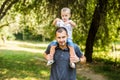 Cute little boy sitting pickaback of his handsome young dad are looking at each other and smiling while resting in the park