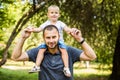 Cute little boy sitting pickaback of his handsome young dad are looking at each other and smiling while resting in the park