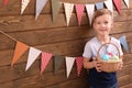 Cute little boy holding basket full of Easter eggs near wooden wall decorated with party pennants