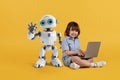 Cute little boy with laptop sitting on floor and smiling to camera, adorable kid robot posing near, orange background Royalty Free Stock Photo