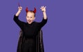 A cute little boy on a purple background, dressed in a black cloak with horns on his head and a trident in his hands, looks at the Royalty Free Stock Photo