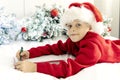 cute little boy in Santa Claus hat writes letter to Santa Claus to get gift lying on bed against background of Christmas Royalty Free Stock Photo