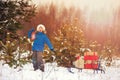 Cute little boy in Santa hat carries a wooden sled with gifts in snowy forest