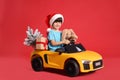 Cute little boy in Santa hat with Christmas tree, bunny toy and gift box driving children`s car on red background Royalty Free Stock Photo