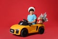 Cute little boy in Santa hat with Christmas tree and gift box driving children`s electric toy car on red background Royalty Free Stock Photo