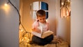 Cute little boy sitting in toy cardboard house at night and reading big fairy tale book. Concept of child education and Royalty Free Stock Photo